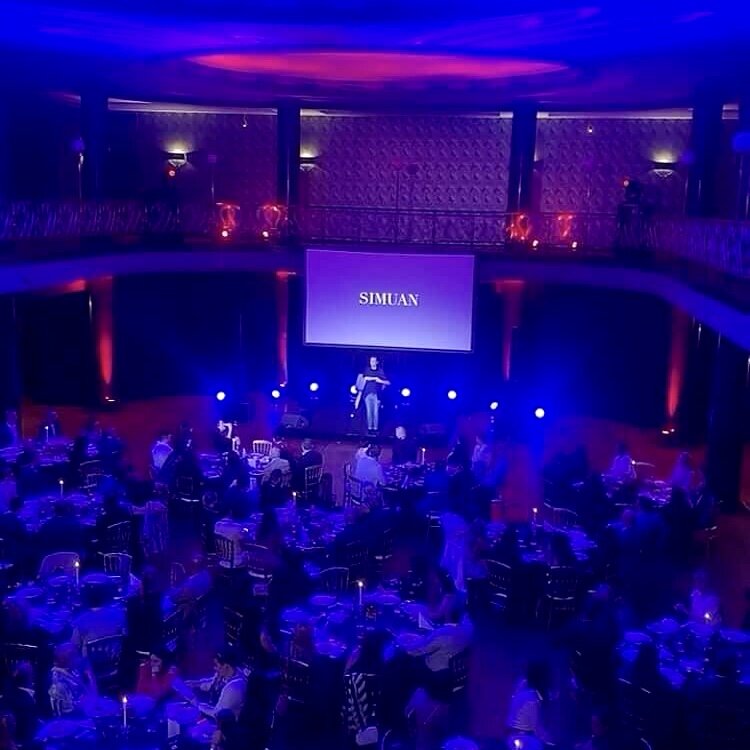 Simuan on stage performing a show. In front of him, several tables.